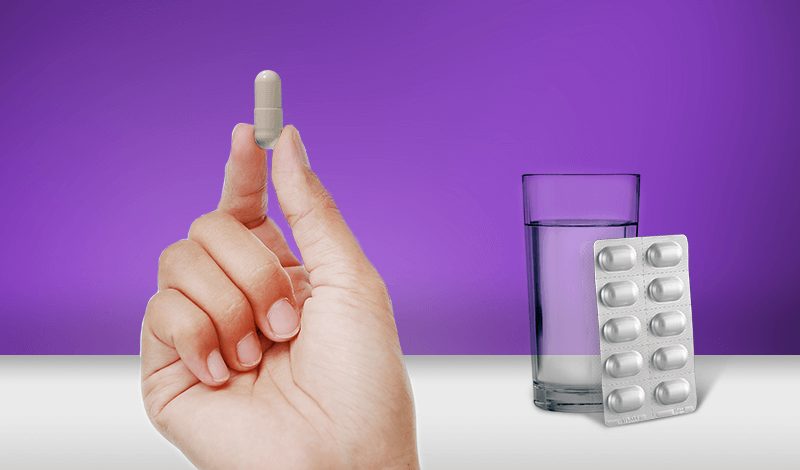A hand holding a probiotic capsule next to a glass of water
